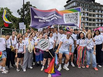 Group of researchers attending the Pride in London march in London, July 2025, wearing white T-shirts printed with the MRS logo in rainbow colours and holding Market Research Society sign, a banner and rainbow flags.